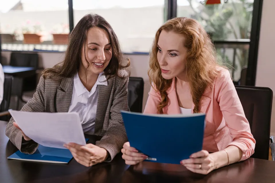 Two businesswomen reviewing documents together in an office setting, emphasizing teamwork and cooperation.