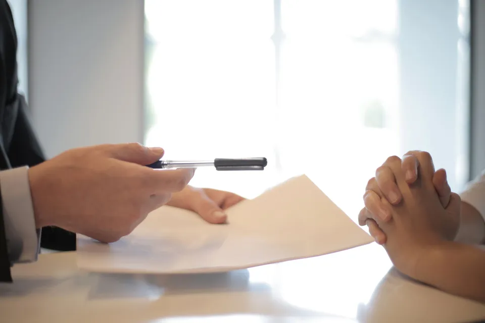 Side view of faceless formal man giving pen and paper to focused female with clenched hands at table on meeting