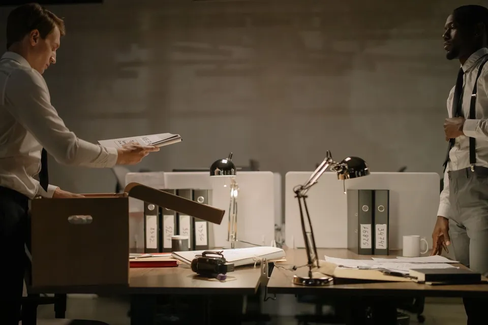 Two office workers sorting documents in a dimly lit workspace with several file binders.