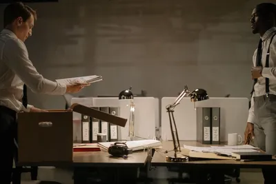 Two office workers sorting documents in a dimly lit workspace with several file binders.